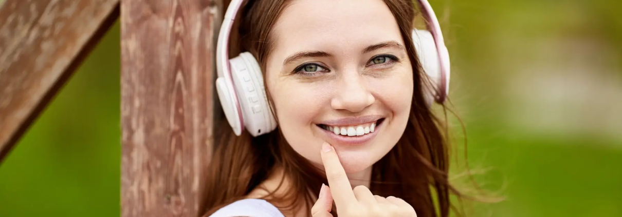 Une jolie femme caucasienne écoute de la musique à l'extérieur à l'aide d'écouteurs sans fil avec un beau sourire et une santé bucco-dentaire parfaite