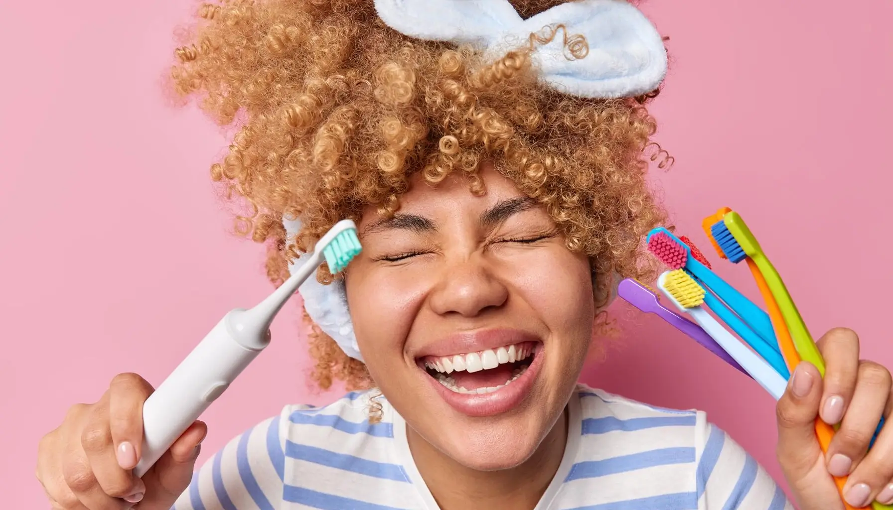 Jeune femme joyeuse avec des cheveux bouclés tenant des brosses à dents, souriant largement, ayant des dents blanches après un nettoyage régulier, portant un t-shirt rayé décontracté, les yeux fermés, isolée sur fond rose.